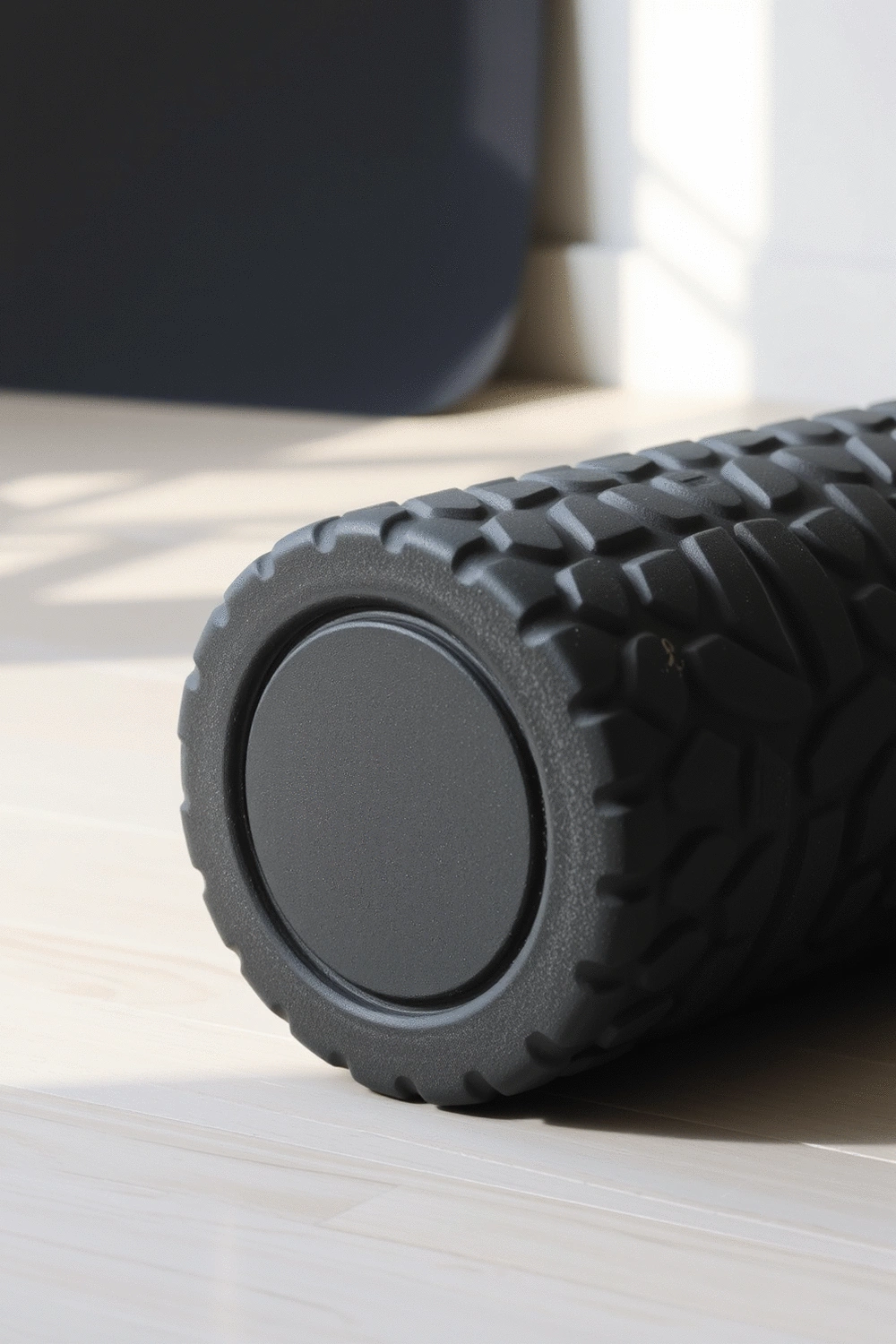 A black foam roller on a clean, light-colored floor, with soft natural light highlighting its texture and cylindrical shape. A yoga mat is partially visible in the background.